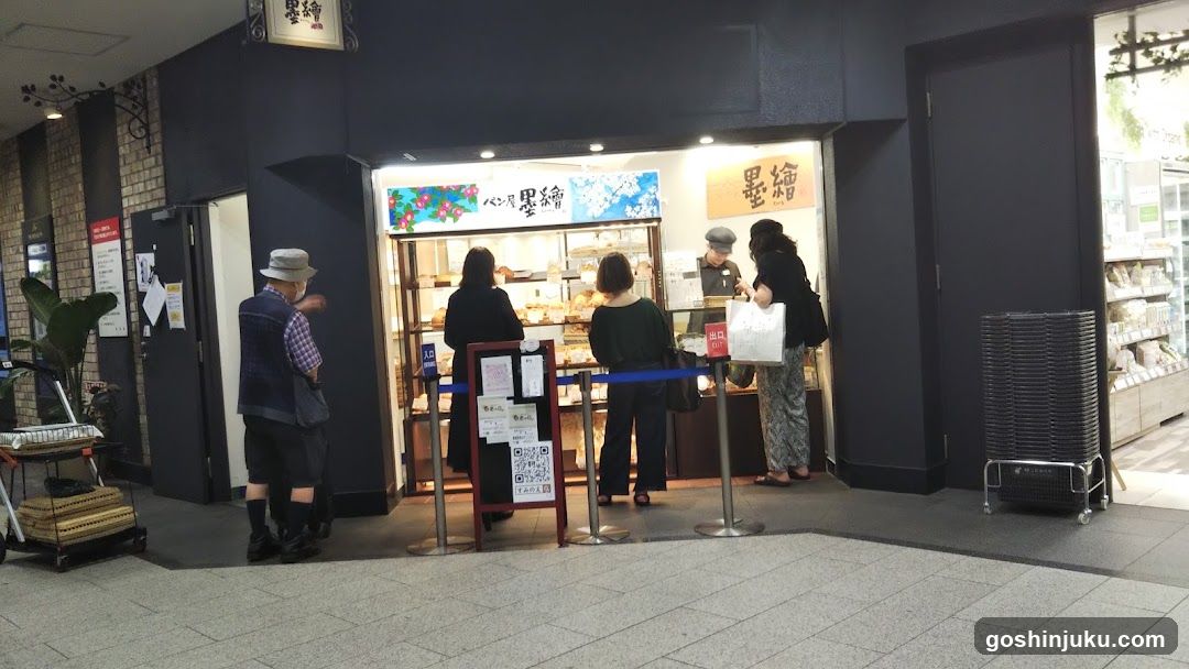 A bakery storefront with visible signage and customers at the counter. The shop has a glass display case showing baked goods, and there are signs above the entrance. A queue barrier is set up outside.