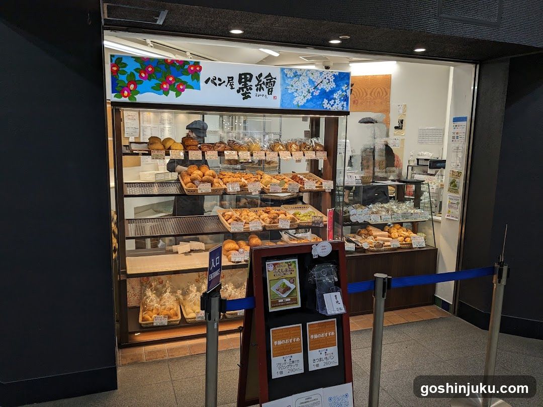 A bakery storefront featuring a glass display case filled with various baked goods like breads, pastries, and cakes. The shop has a sign above the entrance with Japanese characters and floral designs. A menu board stands outside near the entrance.