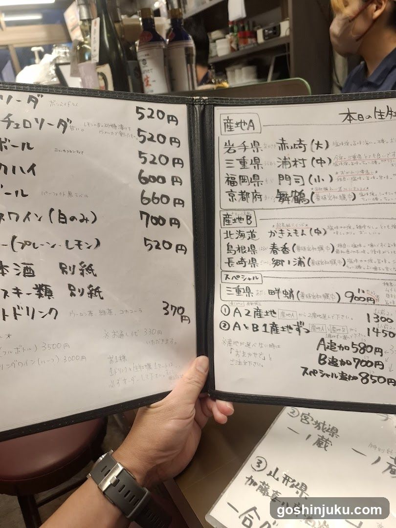 A close-up of a handwritten sake menu in Japanese, held by a person's hand. The menu lists various types of sake with prices and includes sections labeled '产地 A' (Origin A) and '产地 B' (Origin B), detailing different regions like Gifu Prefecture, Mie Prefecture, Fukuoka Prefecture, Kyoto Prefecture, Hokkaido, Aomori Prefecture, Iwate Prefecture, and Tohoku region. It also mentions special offers for orders over 580 yen or 700 yen.
