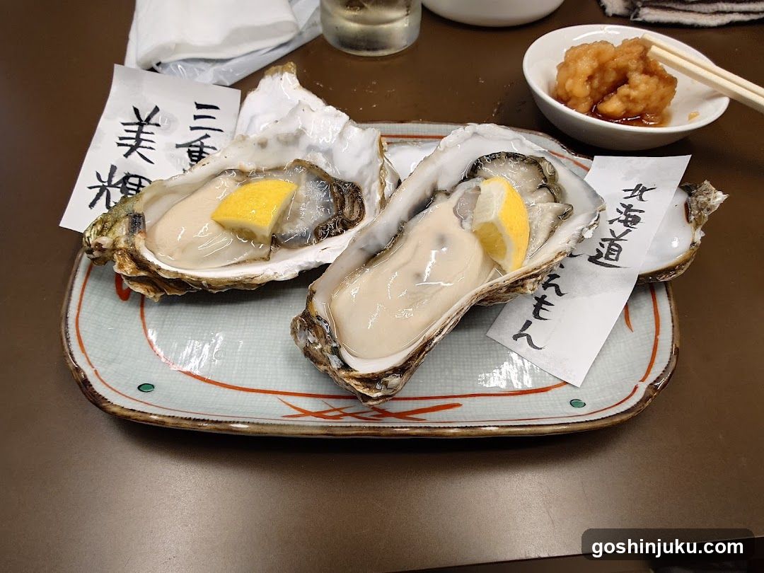 A plate of two oysters, each garnished with a lemon wedge, served on a ceramic dish with handwritten labels in Japanese. A small bowl of sauce and chopsticks are visible nearby.