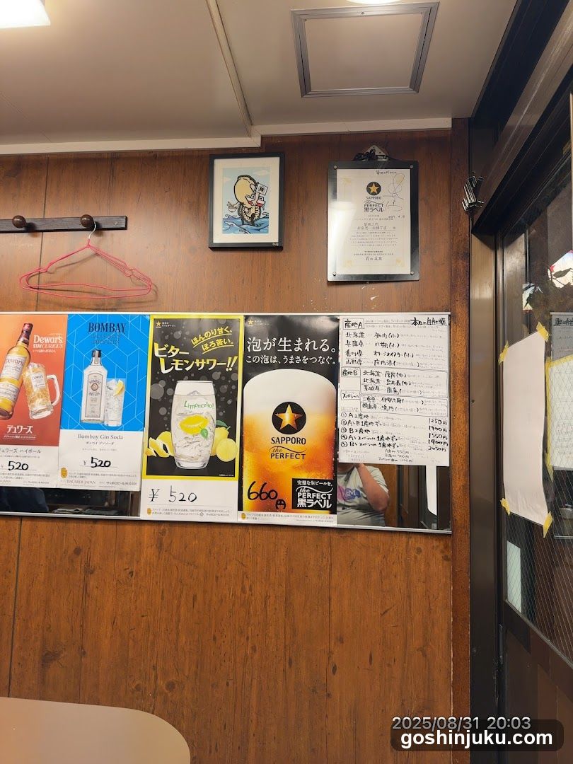A wooden-paneled wall inside a shop displays several promotional posters and a handwritten menu board. Visible items include Bombay Sapphire, Lipton Ice Lemonade, Sapporo Perfect beer, and a list of drinks with prices in yen. A pink hanger hangs on the left, and framed artwork is mounted above.