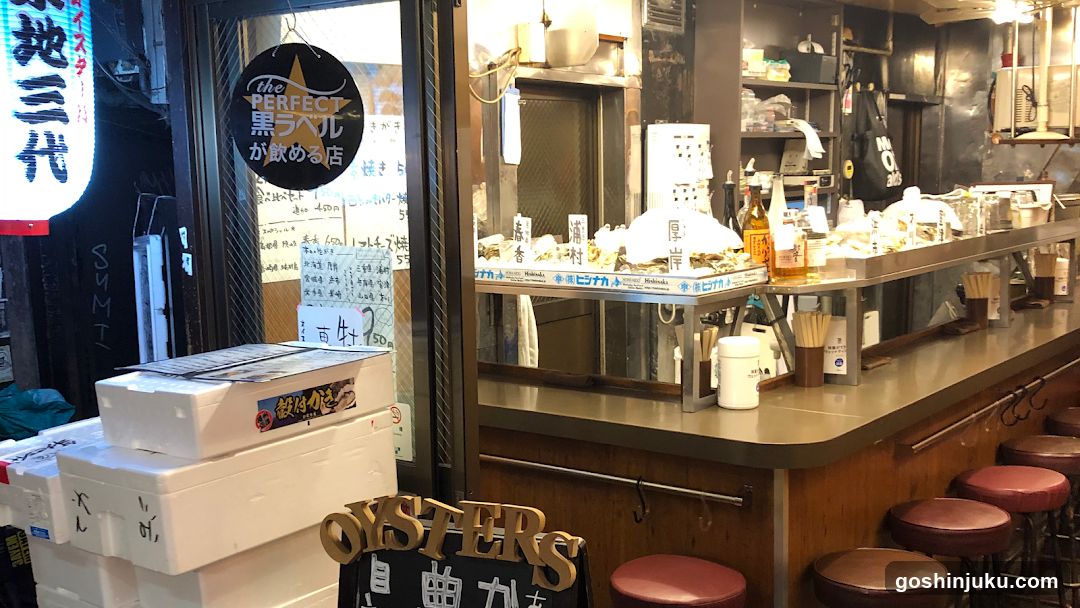 Interior view of an oyster bar with a counter displaying various bottles and containers, stools lined up along the counter, and stacks of white boxes near the entrance. A sign reading 'the PERFECT オパール が飲める店' is visible on the glass door.