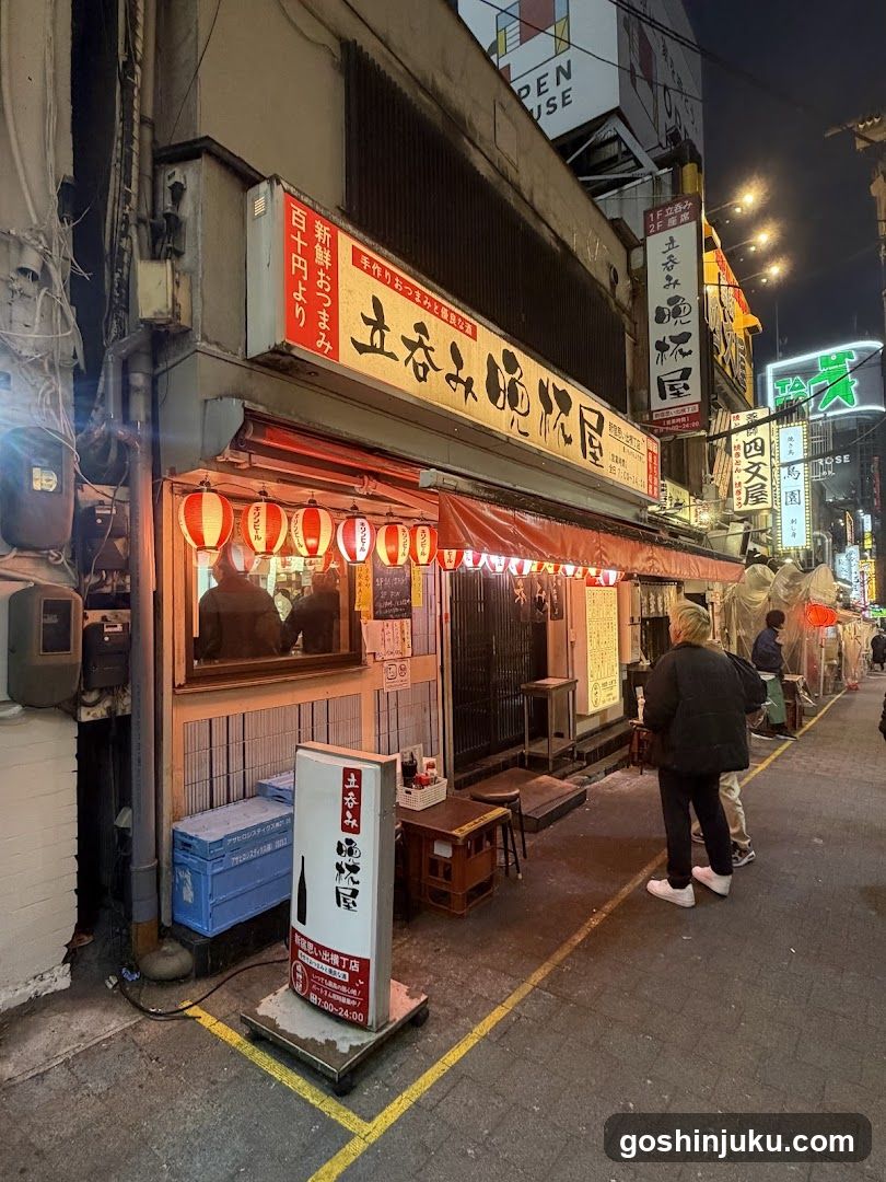 A nighttime view of a narrow urban alleyway featuring a brightly lit storefront with red lanterns hanging above the entrance. A vertical signboard displays Japanese characters and pricing information. Several people are walking along the sidewalk, and additional signage is visible on adjacent buildings in the background.