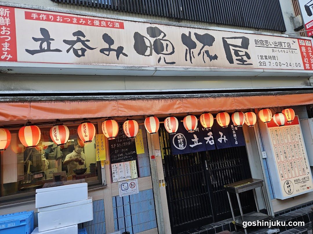 A traditional Japanese shop front with a large signboard displaying '立呑み 屋' (Tachimiya Oshiroiya) and '新宿思い出出店丁店'. The entrance is adorned with red lanterns, and the interior shows staff preparing food behind glass windows. A menu board is visible on the right side of the entrance.