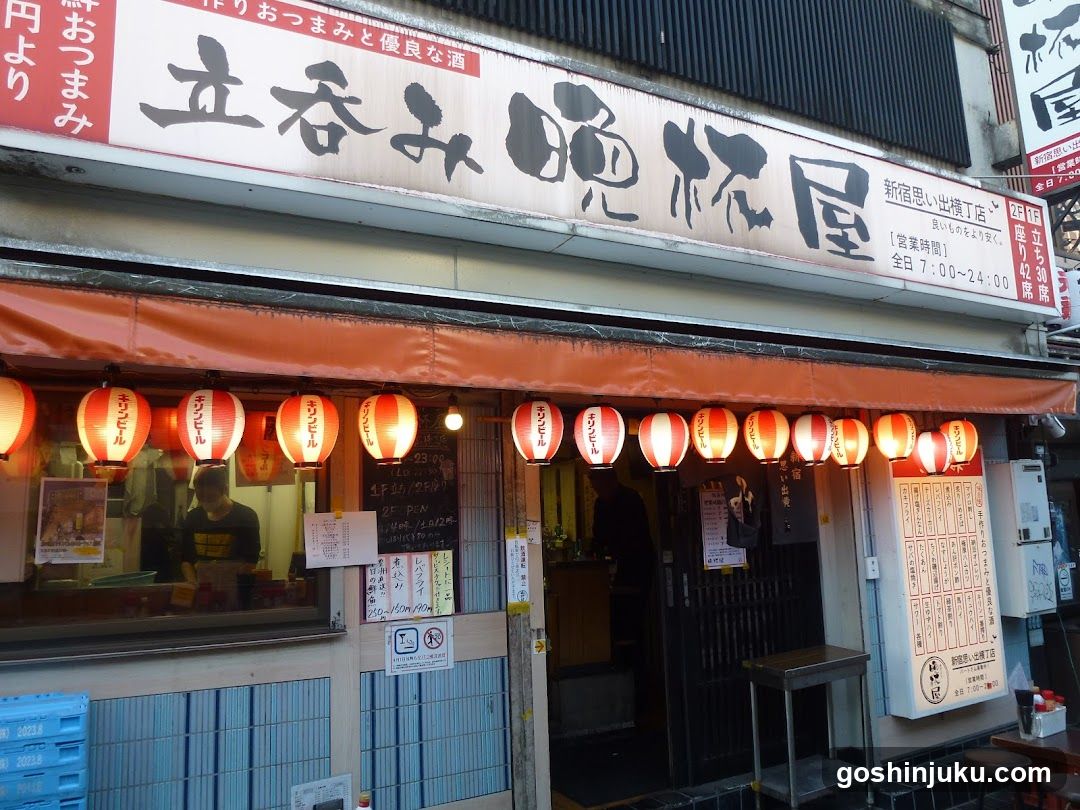A traditional Japanese shop front with a large signboard displaying 'Tachimatsu Sakeya' in bold characters. The entrance is adorned with red lanterns, and the interior is partially visible through glass windows. A menu board is mounted on the wall inside.