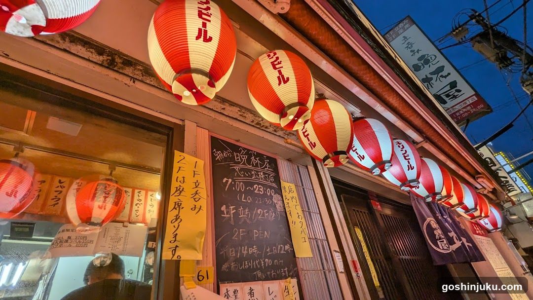 A street-level view of a Japanese restaurant entrance at dusk, featuring red and white paper lanterns hanging above the door. A chalkboard menu lists operating hours (7:00-23:00), seating options (1F/2F), and mentions 'Izakaya' style dining. Yellow signs advertise special promotions like free entry or discounts. The building facade shows traditional signage in Japanese characters, with visible power lines overhead against a dark blue sky.
