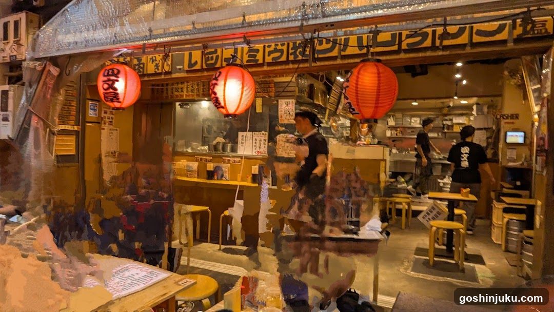 A dimly lit Japanese restaurant storefront with red lanterns hanging above the entrance. The facade features a signboard with Japanese characters and English text 'SHISHIMO'. Inside, staff members are visible near the counter, and tables with stools are arranged in the dining area.