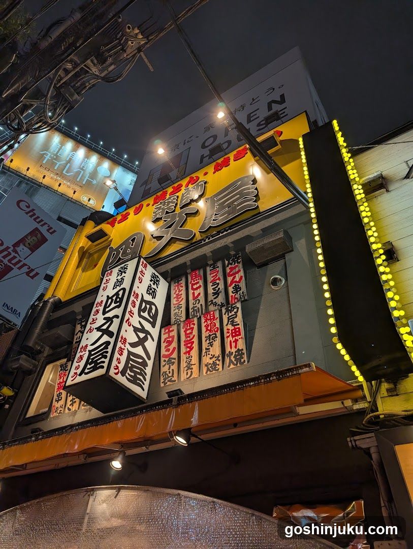A brightly lit shop front at night, featuring a yellow building facade with large Japanese characters on the signboard. Vertical white banners list menu items in red and black text. String lights line the right side of the entrance. Overhead wires and adjacent billboards are visible above.