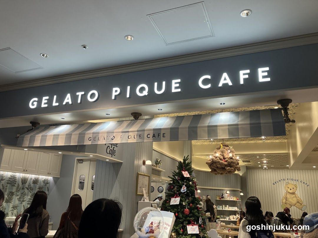 Storefront with illuminated signage reading 'GELATO PIQUE CAFE' and striped awning. Interior visible through open entrance, featuring Christmas tree, hanging decoration, and customers seated at tables.