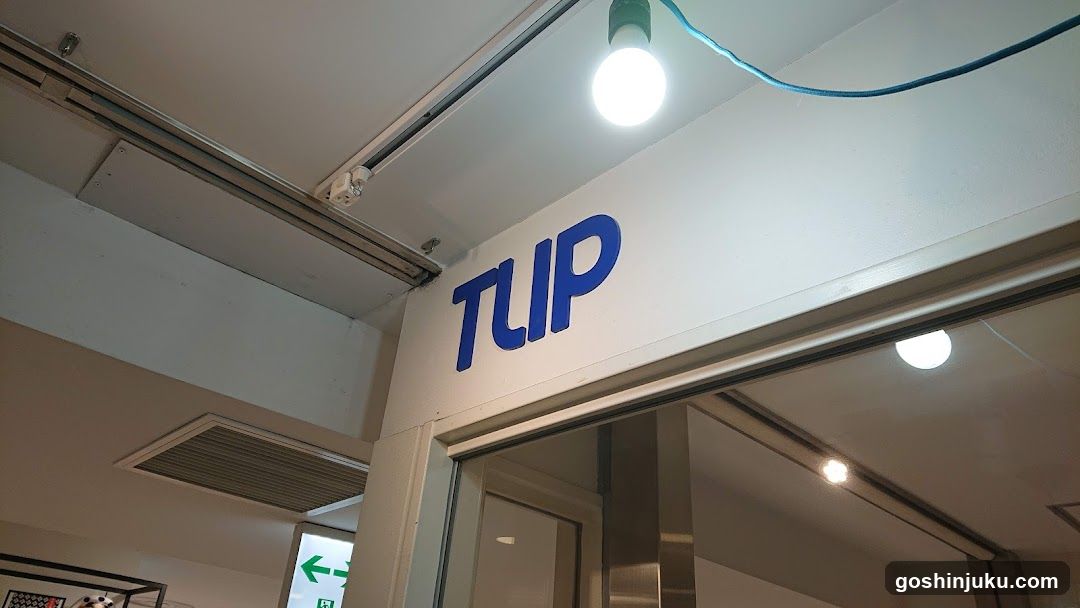 A shop entrance with the word 'TLUP' in blue letters on a white wall above the door. A bright light bulb hangs from the ceiling, and there is a glass door leading inside.