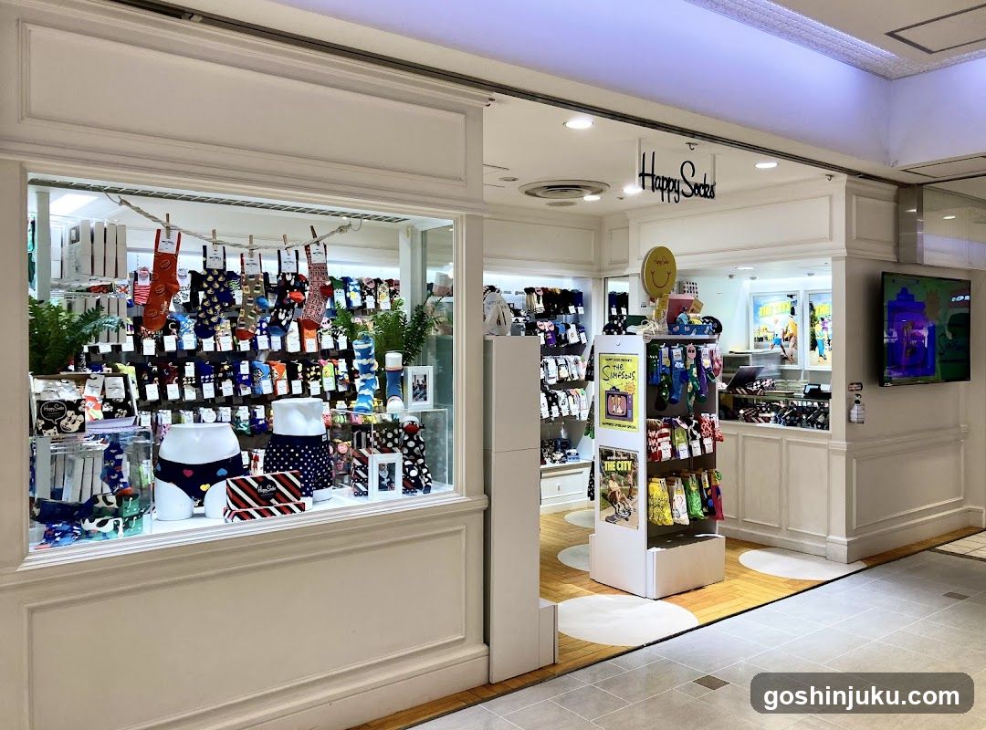 A brightly lit storefront with a large window display showcasing colorful socks and underwear on mannequins. The shop sign reads 'Happy Socks' above the entrance. Inside, shelves are stocked with various sock designs, and promotional posters are visible near the counter.