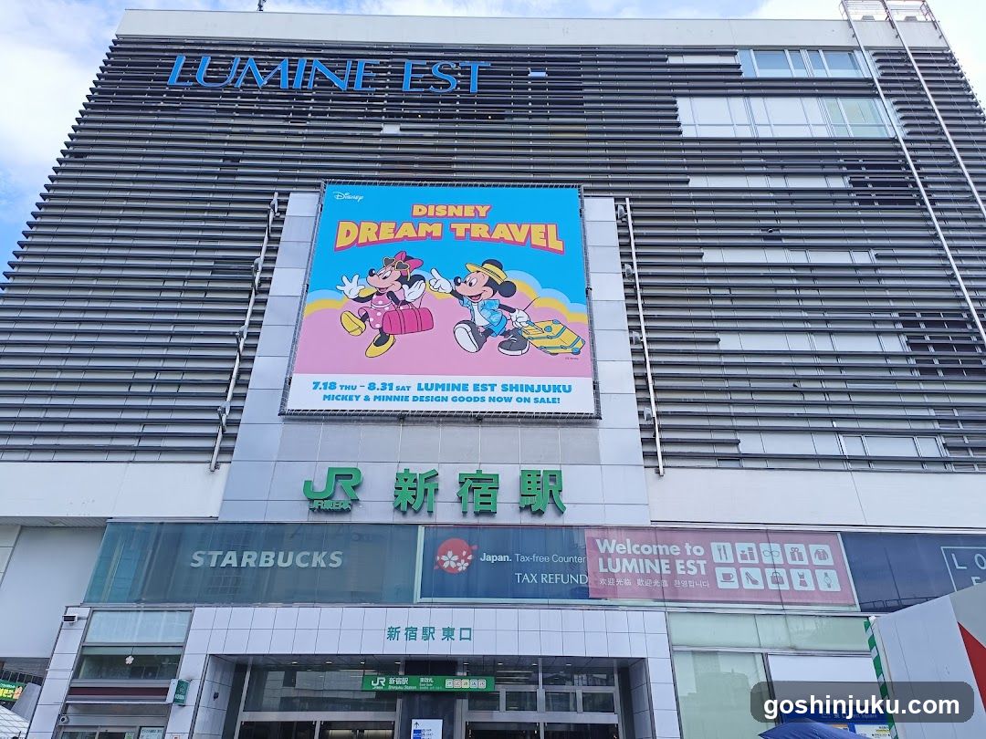 Exterior facade of Lumine Est Shinjuku station building featuring a large Disney Dream Travel advertisement banner, JR logo and Japanese characters, Starbucks signage, tax refund counter sign, and entrance doors with directional signs.