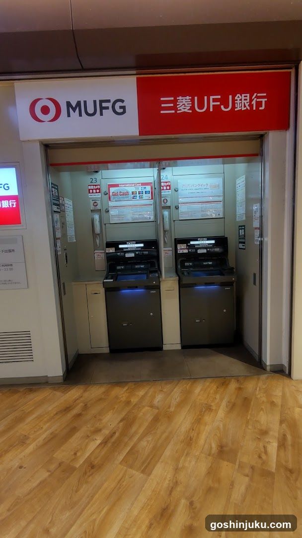 A photo of the entrance to a MUFG (Mitsubishi UFJ Financial Group) bank area, featuring two ATMs under a red and white sign with Japanese characters '三菱UFJ銀行'. The floor is wooden, and there are informational posters on the walls.