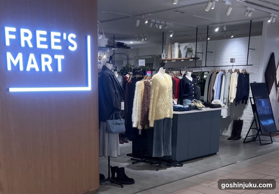 Interior view of a clothing store with illuminated 'FREE'S MART' sign on wooden wall, mannequins displaying winter coats and sweaters, hanging garments, display table with folded clothes, white brick walls, track lighting, and digital screen stand.