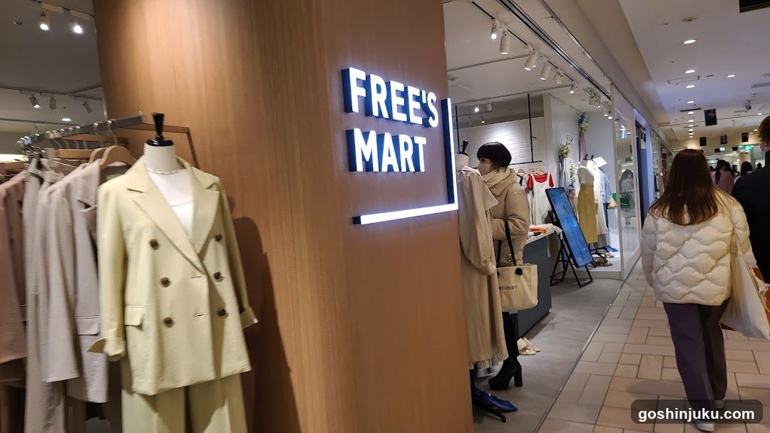 A storefront of Free's Mart, a clothing store in a shopping mall. The sign is illuminated with blue LED lights on the wooden facade. Mannequins display beige and cream-colored outfits. A woman browses inside while another walks by.