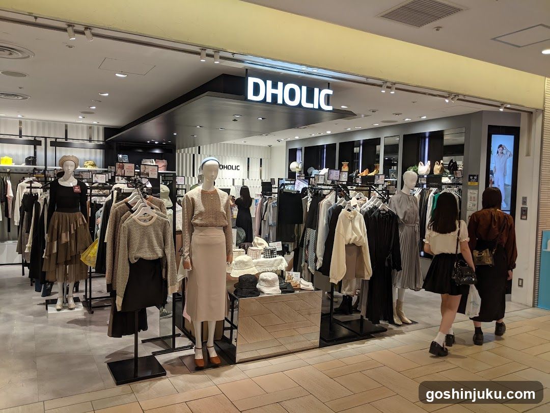 Interior view of the Dholic clothing store, showcasing mannequins dressed in various outfits including skirts and tops. The store features a modern design with racks of clothes, accessories on display tables, and customers browsing near the entrance.