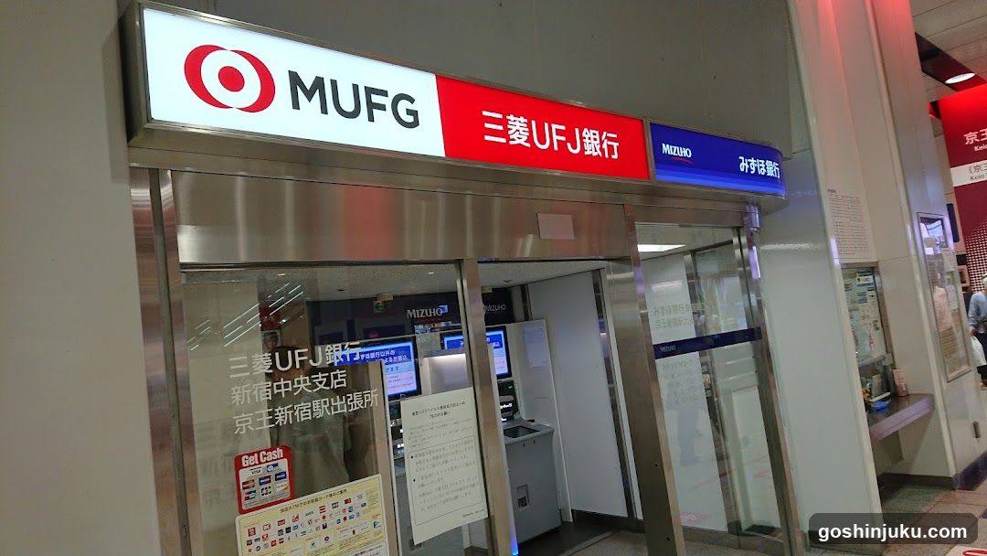 A photo of the physical exterior entrance/storefront facade of MUFG Mizuho Bank, specifically the Shinjuku Chuo branch located at King Seiko Station Exit. The signage above the entrance displays 'MUFG' with its logo and '三菱UFJ銀行' (Mizuho UFJ Bank) in Japanese characters alongside English text. A blue sign indicates 'みずほ銀行' (Mizuho Bank). Glass doors lead into the bank interior where ATMs are visible inside.