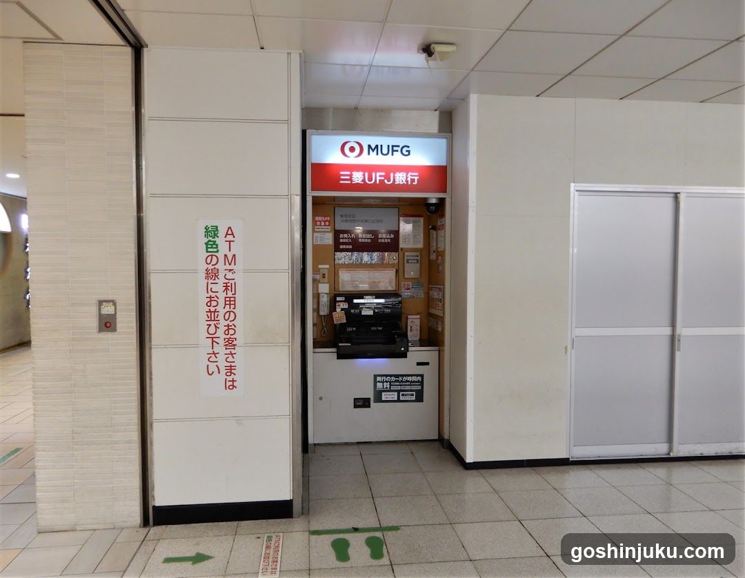 A MUFG (Mitsubishi UFJ Financial Group) ATM kiosk located in a public indoor space, likely a train station or shopping mall. The kiosk features the MUFG logo and name on its signage above the machine. A sign next to it instructs customers to queue behind those using green lines. The floor has directional arrows and footprints indicating queuing order.