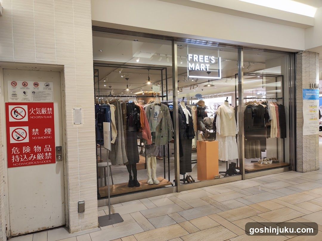 A clothing store named 'FREE'S MART' with glass windows displaying mannequins and racks of clothes. A red sign on the left door indicates no open flame, no smoking, and no dangerous goods.