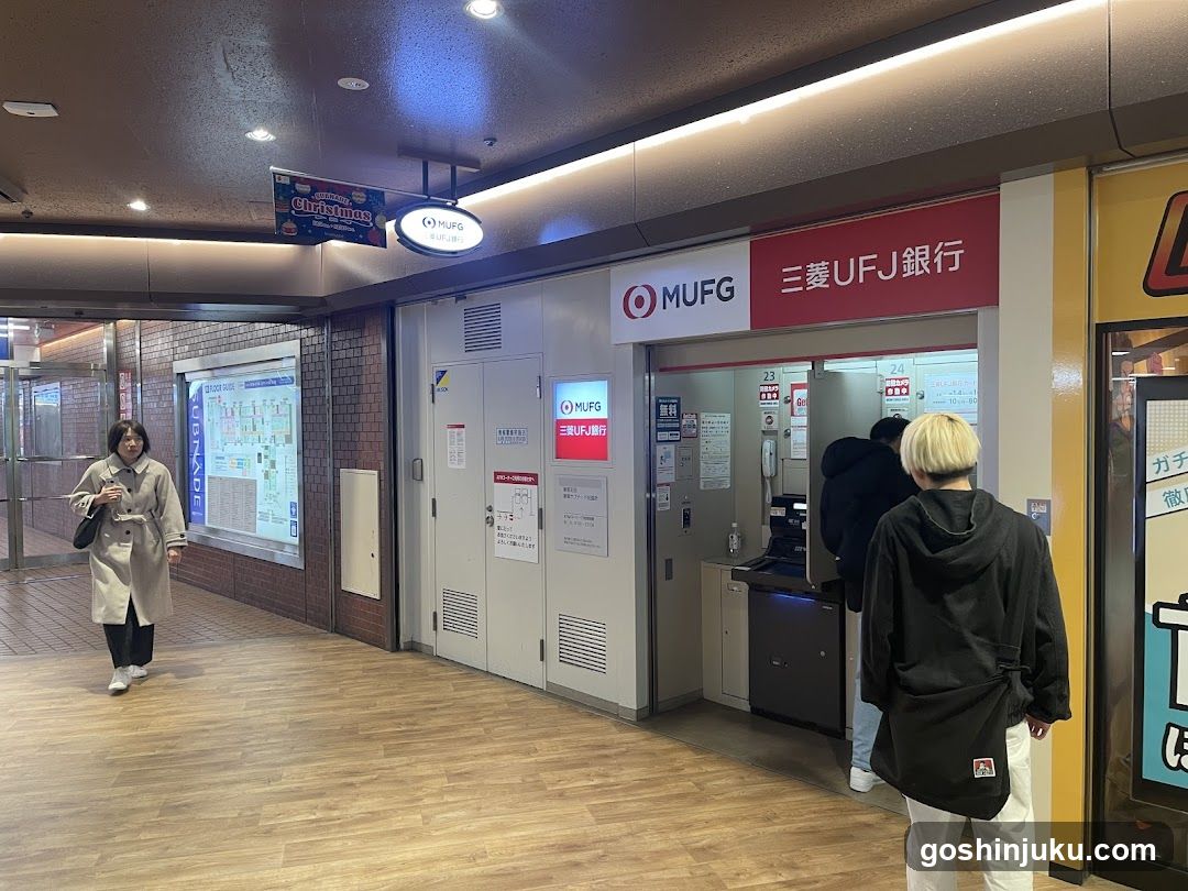 A photo of the physical exterior entrance to a MUFG (Mitsubishi UFJ Financial Group) bank branch, featuring signage with 'MUFG' and '三菱UFJ銀行' visible on the building facade. A person in black stands near the entrance while another walks by.