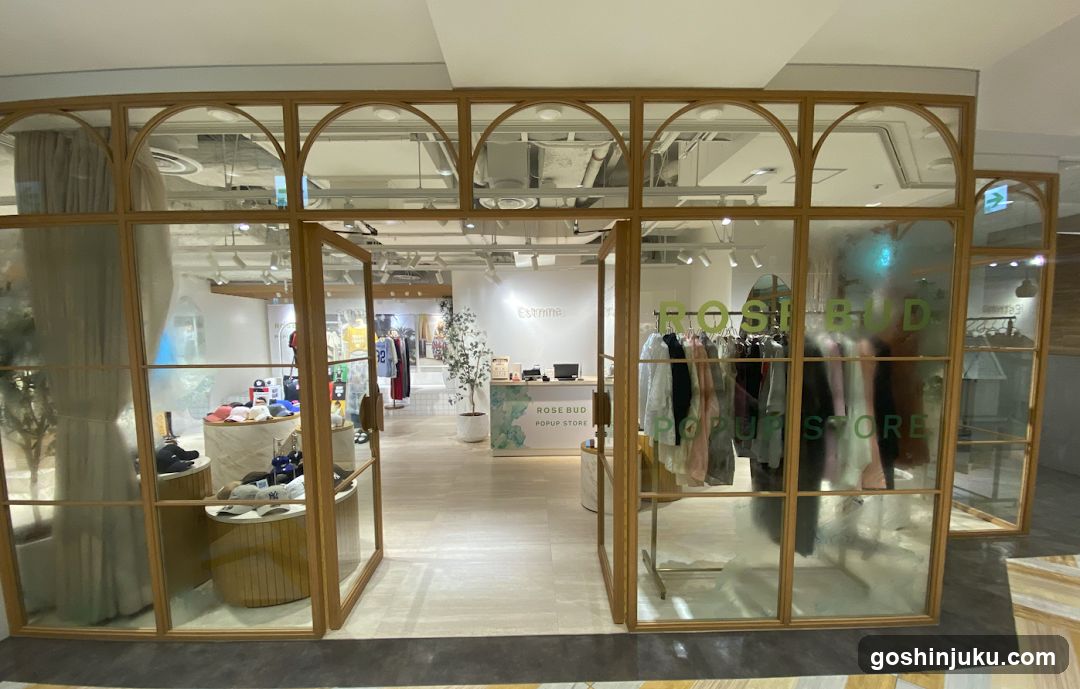 A modern retail storefront with arched glass windows and wooden frames. The entrance features double doors, one slightly ajar, revealing an interior display of clothing racks and merchandise. Signage on the window reads 'ROSE BUD POPUP STORE' in green lettering. Inside, a counter displays promotional materials and products are arranged neatly under bright lighting.