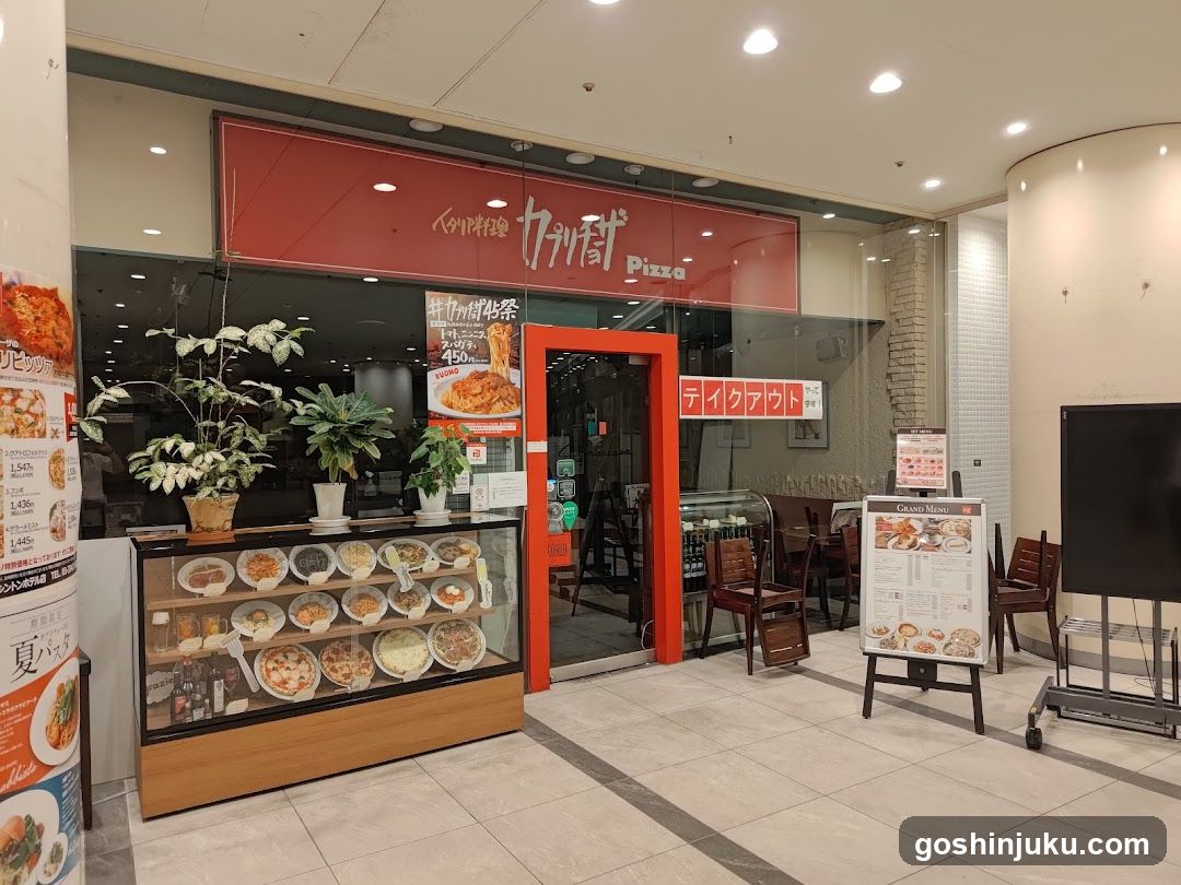 A storefront with a red sign displaying 'Kakugawa Pizza' in Japanese and English. The entrance has an orange frame, and there are potted plants outside. A glass display case shows various food items inside. There is also a menu board on the right side of the image.