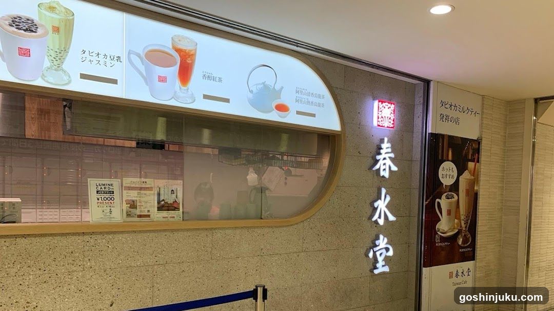 The image shows the storefront of a tea shop named '春水堂' (Haruizuya). The facade features illuminated vertical signage with the shop name in Japanese characters. Above the entrance, there is a menu board displaying various beverages including milk teas and iced drinks. A poster on the right side advertises additional products like bubble tea.