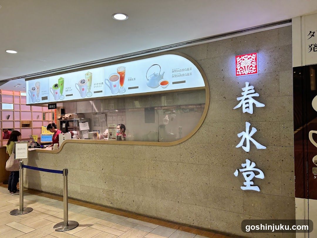 A shop front with a large illuminated sign reading '春水堂' (Shunsui-do) on the right side of the facade. The counter area features a curved glass window displaying various drink options above it, including green tea-based beverages and milk teas. Staff members are visible behind the counter attending to customers.