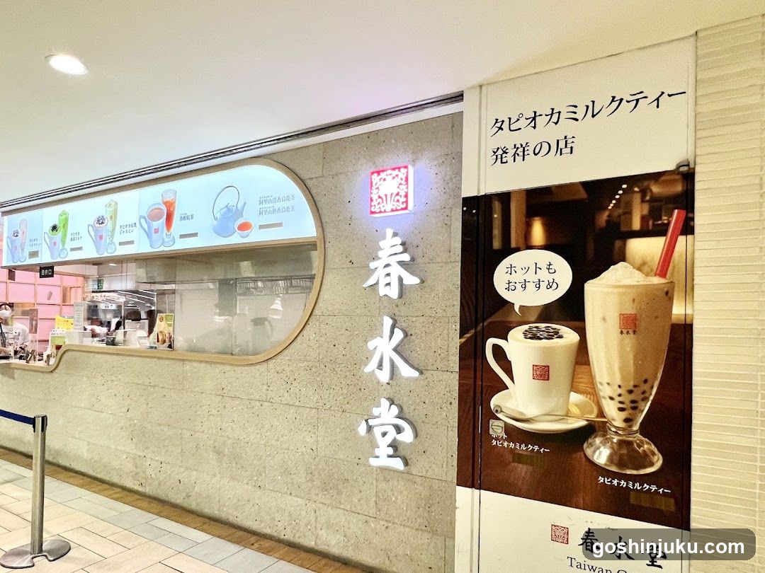 The image shows the storefront of a bubble tea shop with illuminated signage reading '春水堂' (Chun Shui Tang). A large poster to the right advertises their signature milk tea and boba pearls, featuring images of both hot and cold drinks. The shop has a modern design with a concrete facade and a display window showcasing various drink options.