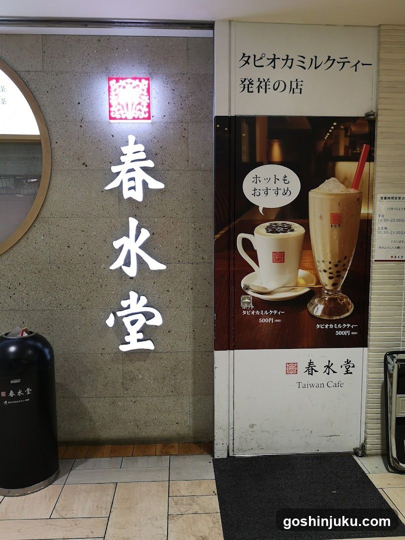 A storefront entrance featuring illuminated vertical Japanese characters '春水堂' (Haruizuya Dojo) on a textured wall, alongside a promotional poster advertising bubble milk tea drinks with prices listed in yen.