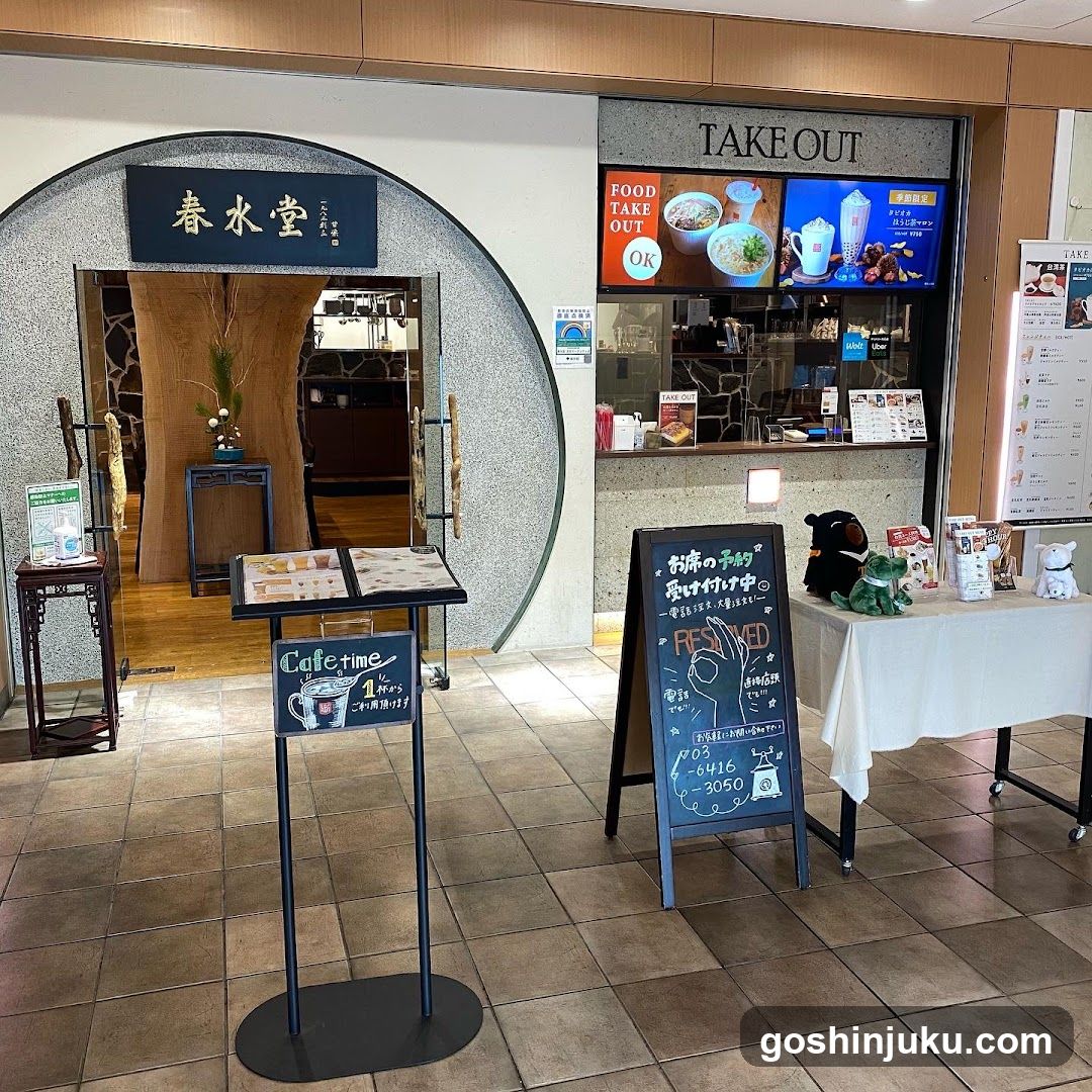 A storefront with a circular entrance and signage reading '春水堂' above the door. To the right, there is a TAKE OUT window displaying food items on screens. Outside, there are chalkboard signs advertising cafe time and takeout options, along with a table covered in white cloth holding various products and plush toys.
