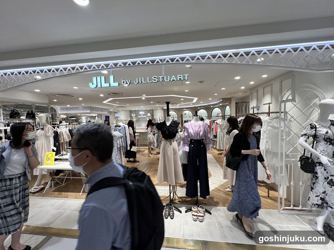 The image shows the storefront of a clothing store named 'JILL by JILLSTUART'. The sign is prominently displayed above the entrance with illuminated lettering. Inside, there are mannequins dressed in various outfits, including dresses and skirts. Shoppers can be seen browsing the merchandise. The interior features modern lighting and wooden flooring.