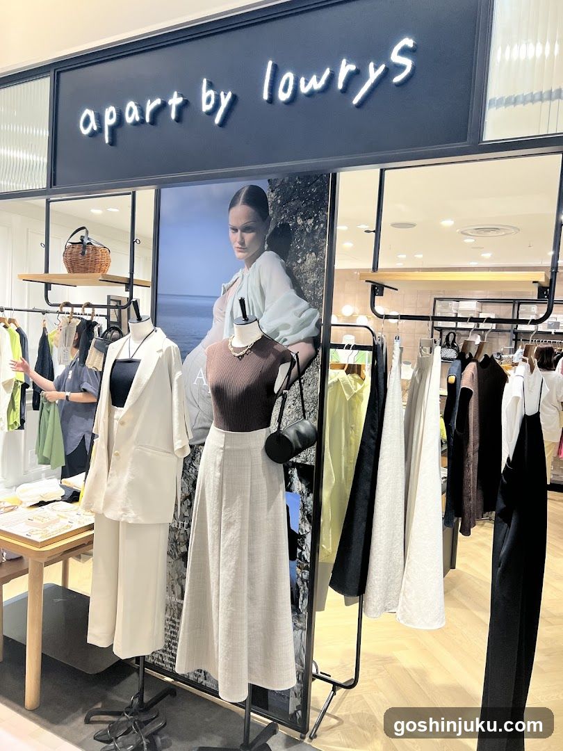 The image shows the storefront of a clothing store named 'apart by lowryS'. The sign is mounted above the entrance with illuminated lettering. Inside, mannequins display fashion outfits including a white suit and a brown top with a long skirt. Clothing racks are visible on both sides, displaying various garments in neutral tones. A large poster featuring a model stands behind the central display. Shelves hold accessories like baskets. The interior has light wood flooring and modern lighting.