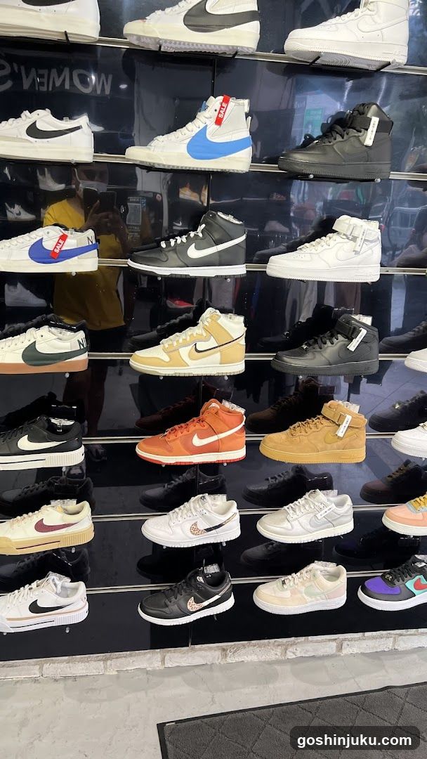 A wall display of various Nike sneakers, including high-tops and low-tops in multiple colors like white, black, blue, orange, and beige. Some shoes have tags attached. The background shows reflections of people taking photos.