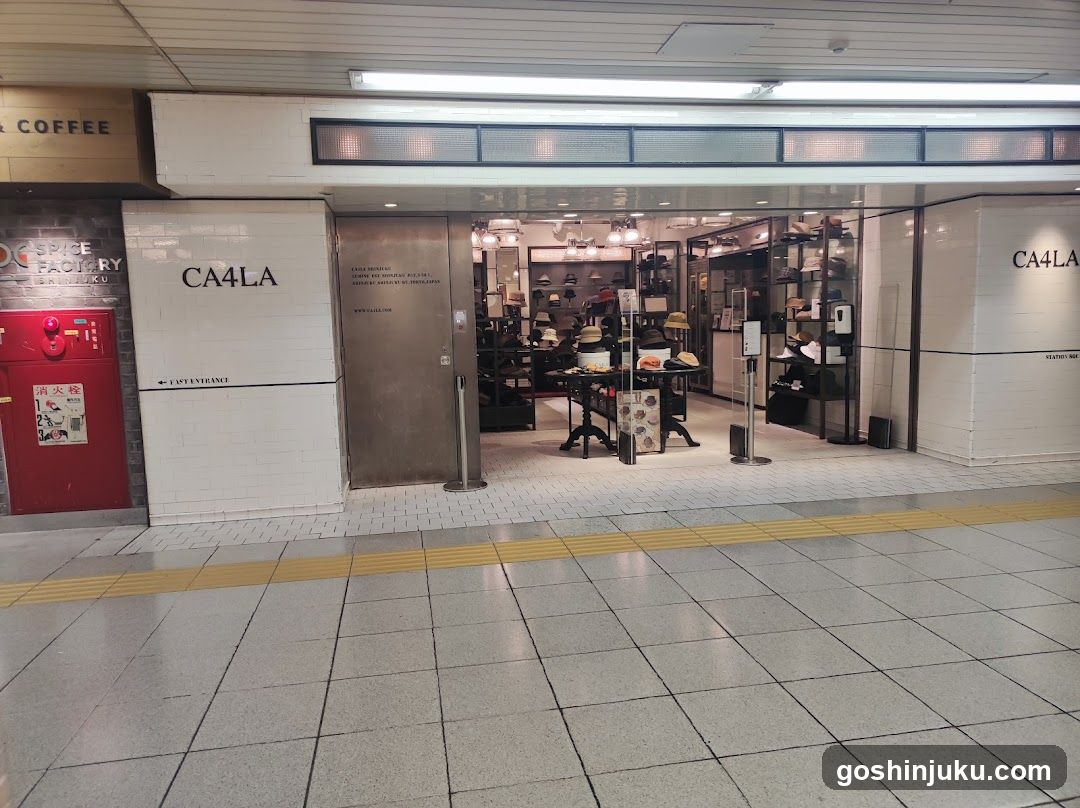 A shop front with visible signage reading 'CA4LA' on white tiled walls. The entrance is open, revealing an interior display of hats and accessories. A red fire alarm box is mounted to the left, and a yellow tactile paving strip runs along the floor in front.