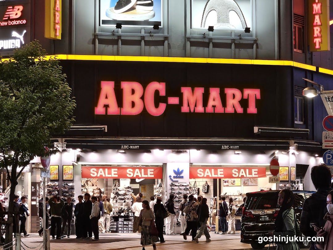 A brightly lit ABC-MART storefront with large red signage on a black facade. The entrance features multiple 'SALE' banners above glass doors and windows displaying shoes and apparel. Pedestrians walk along the sidewalk in front, and a tree is visible to the left. Adjacent signs include Puma and New Balance logos.