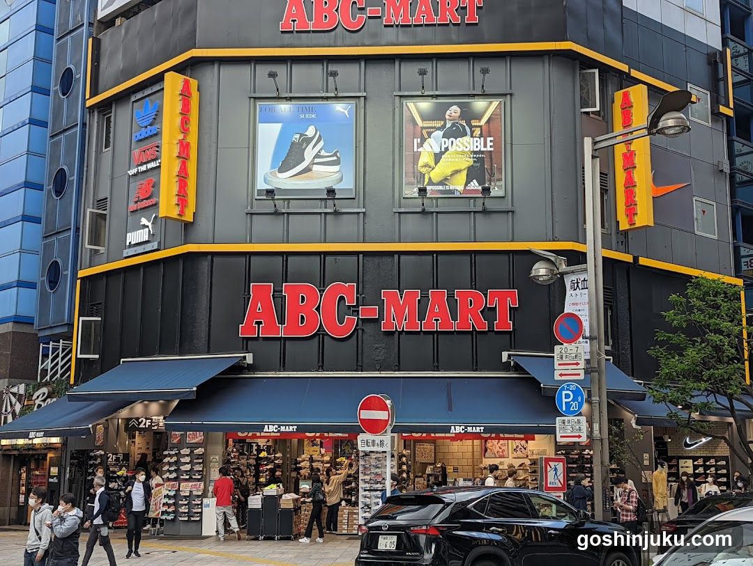 A street-level view of an ABC-MART store with prominent red signage on the facade and awnings. Large windows display shoe advertisements, including a sneaker and a model in yellow. Pedestrians walk by, and a black car is parked nearby.