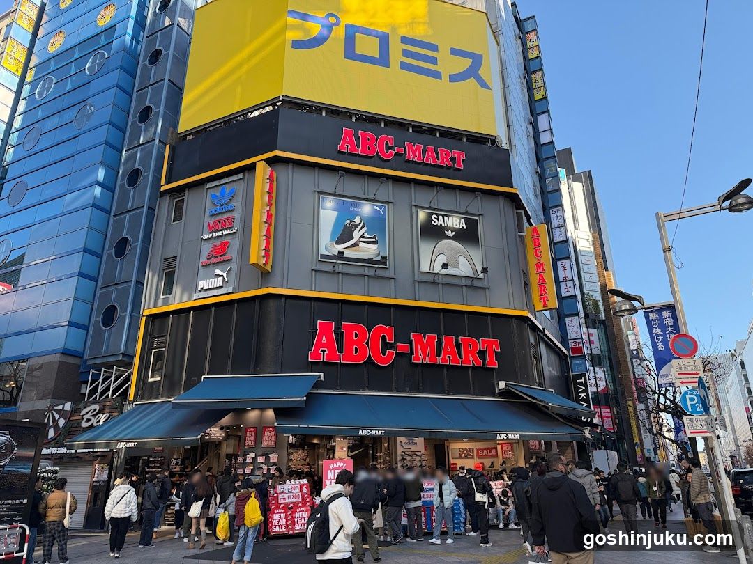 A busy street corner featuring an ABC-MART store with prominent signage on the building facade. The storefront has blue awnings and displays large shoe advertisements above the entrance. Pedestrians are walking along the sidewalk, and various signs and banners are visible around the area.