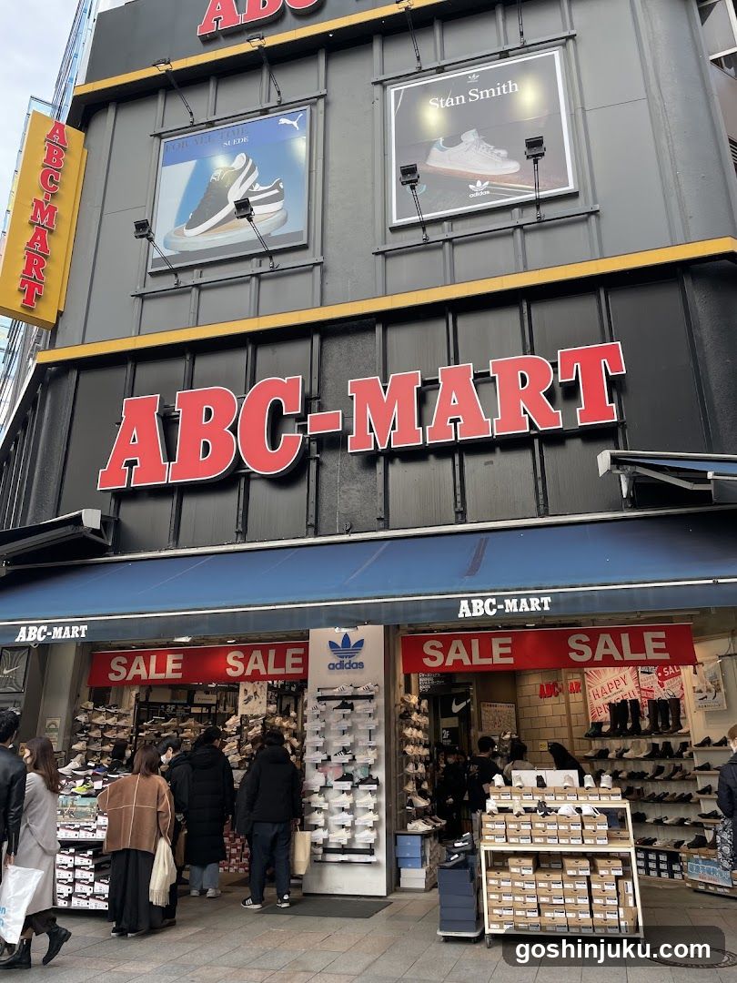 The image shows the exterior of an ABC-MART store, a well-known shoe retailer in Japan. The building facade features large red signage with 'ABC-MART' prominently displayed. Above the entrance, there are advertisements for Puma and Stan Smith sneakers. The storefront has blue awnings with 'ABC-MART' written on them. Several 'SALE' signs are visible above the entrances to different sections of the store. Shelves filled with shoes line both sides of the entrance, and customers can be seen browsing inside.