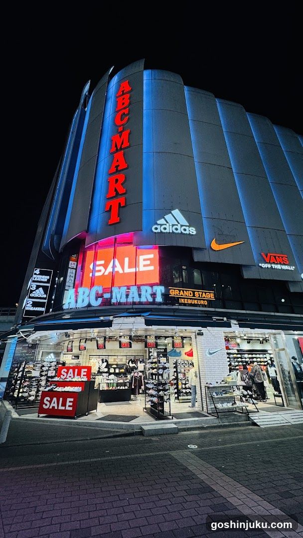 Night view of ABC-MART building exterior with illuminated signage, including 'SALE' banners and brand logos like Adidas, Nike, Vans. Store entrance displays shoes and apparel on racks.