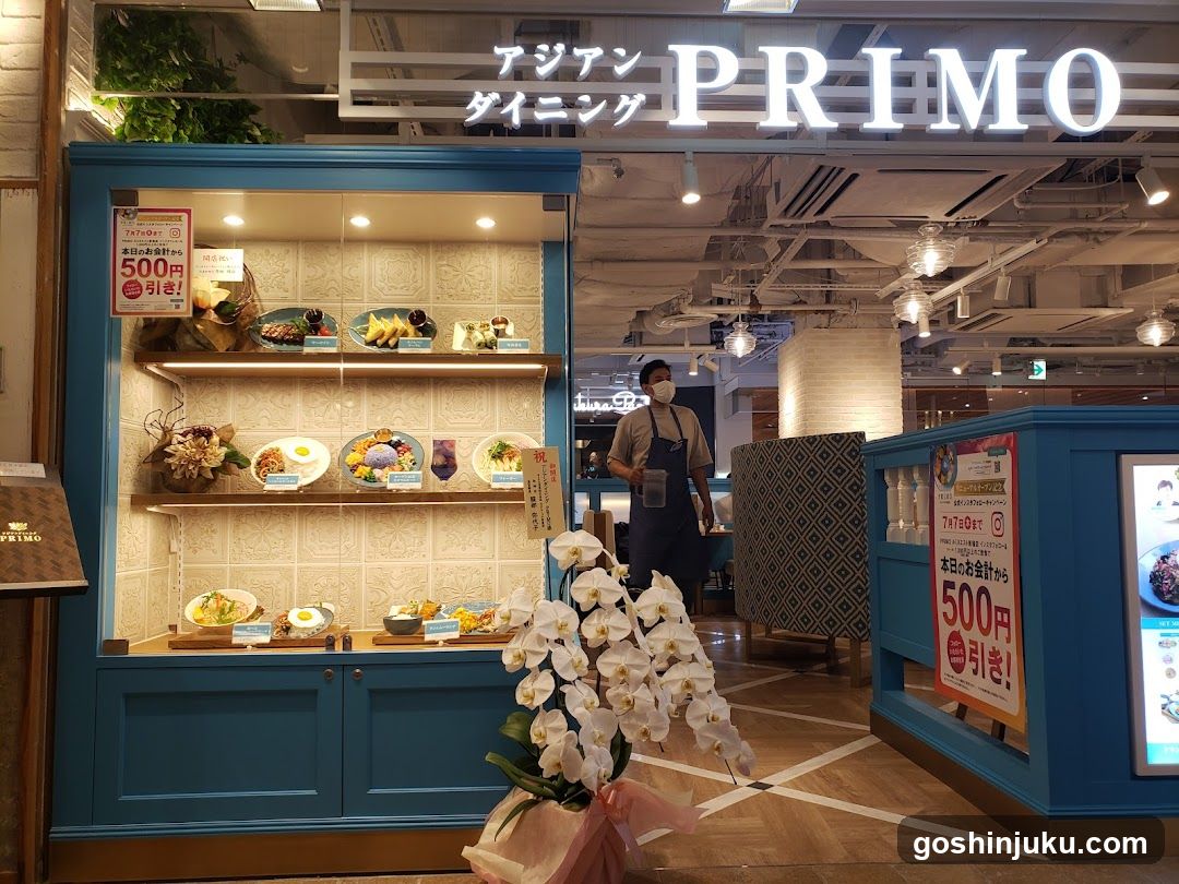 A brightly lit storefront with blue paneling and white tiled backsplash displaying food samples on shelves. A sign above reads 'アジアン ダイニング PRIMO'. A staff member in an apron stands near the entrance. Decorative white orchids are placed at the base of the display. Posters advertise a 500 yen discount.