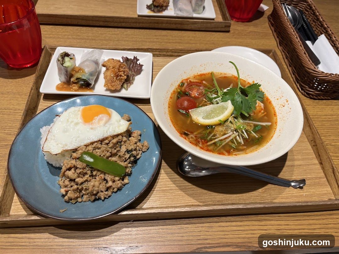 A wooden tray holding a variety of Thai dishes including Tom Yum soup, fried rice with egg and green chili, spring rolls, and crispy chicken pieces.