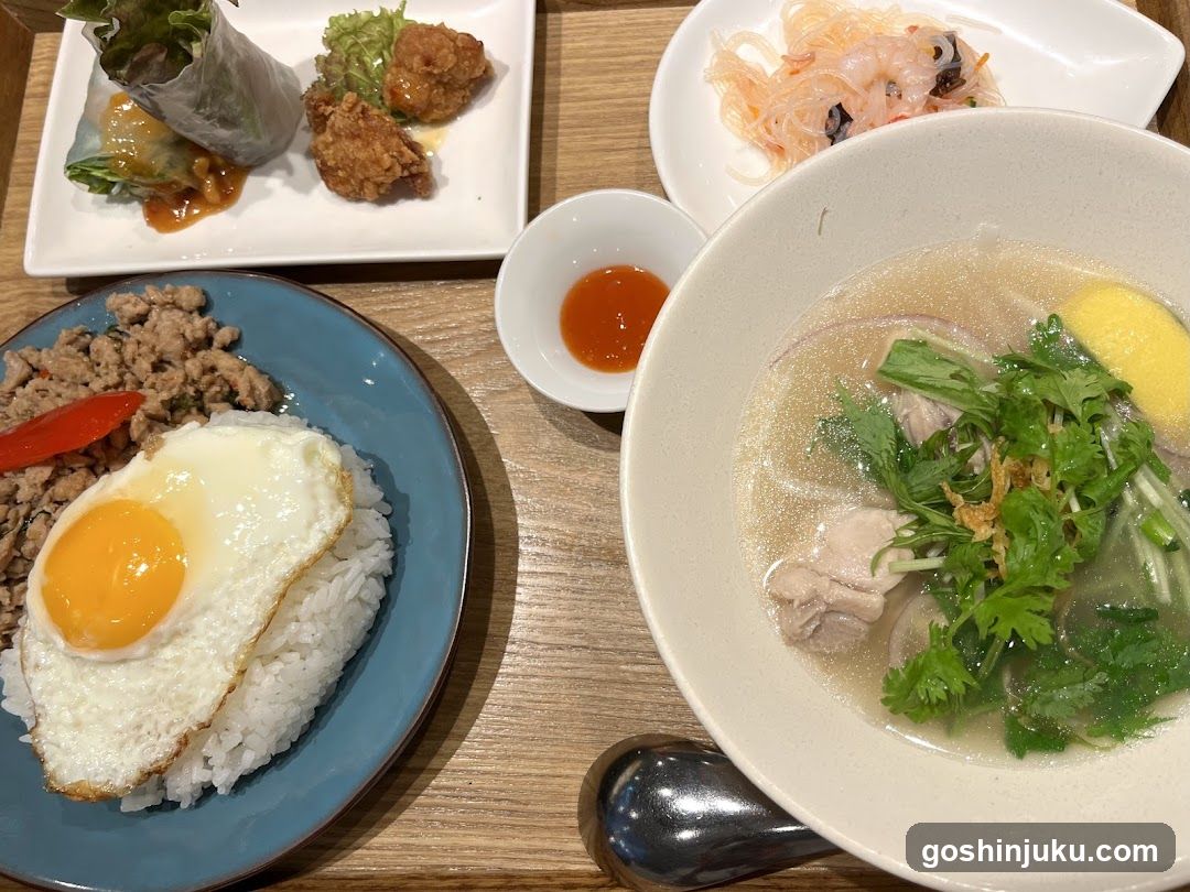 A table setting featuring a bowl of noodle soup with egg and herbs, a plate of rice topped with fried egg and minced meat, spring rolls with dipping sauce, shrimp salad, and a small dish of red chili sauce.