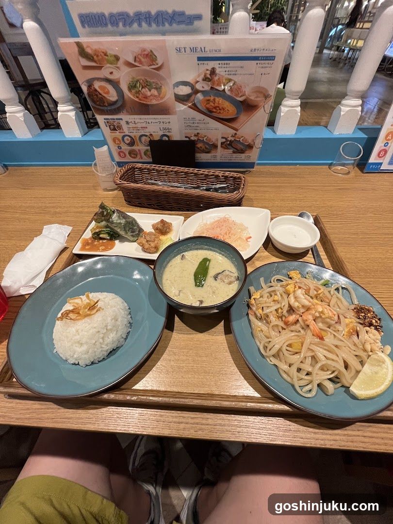 A wooden table set with a tray containing rice, curry soup, stir-fried noodles, and side dishes including pickled vegetables and shrimp. A menu board in the background displays various meal options with images.