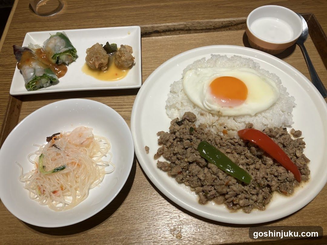 A wooden table displays a Thai-inspired meal consisting of a large plate with white rice topped by a sunny-side-up egg and minced meat mixed with green and red peppers. Accompanying sides include glass noodles with shrimp, spring rolls with sauce, two meatballs in gravy, and a small bowl of broth or soup.