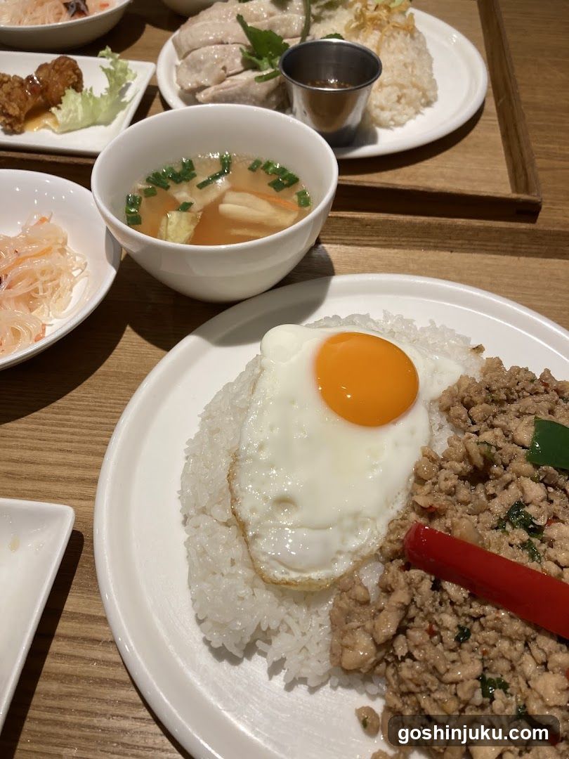 A close-up of a meal featuring fried rice topped with a sunny-side-up egg, accompanied by minced pork stir-fry, a bowl of clear soup with tofu and green onions, and side dishes including pickled vegetables and chicken pieces.