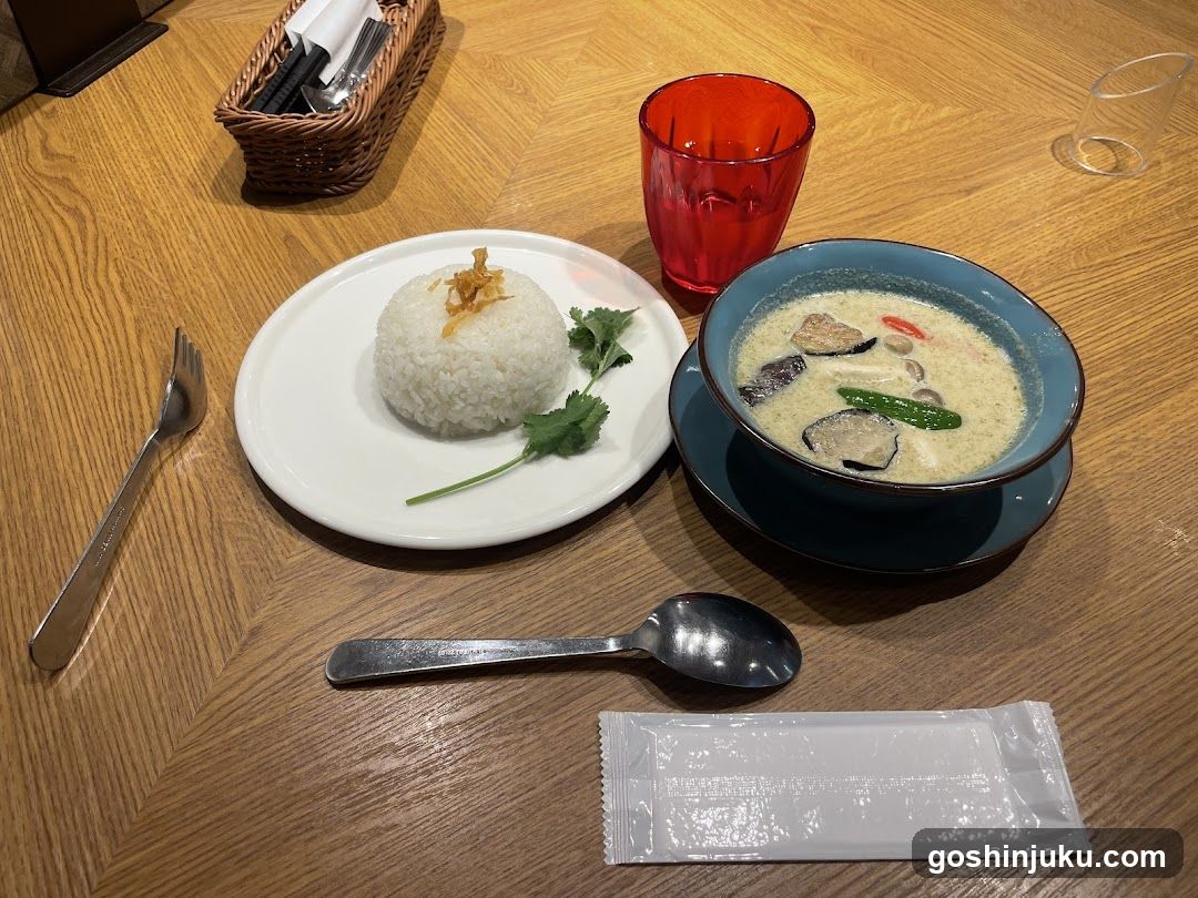 A table setting featuring a bowl of creamy coconut curry with eggplant, green beans, and red peppers; a plate of white rice topped with fried shallots and garnished with cilantro; a red glass; silverware wrapped in paper; and a small basket holding cutlery on a wooden surface.