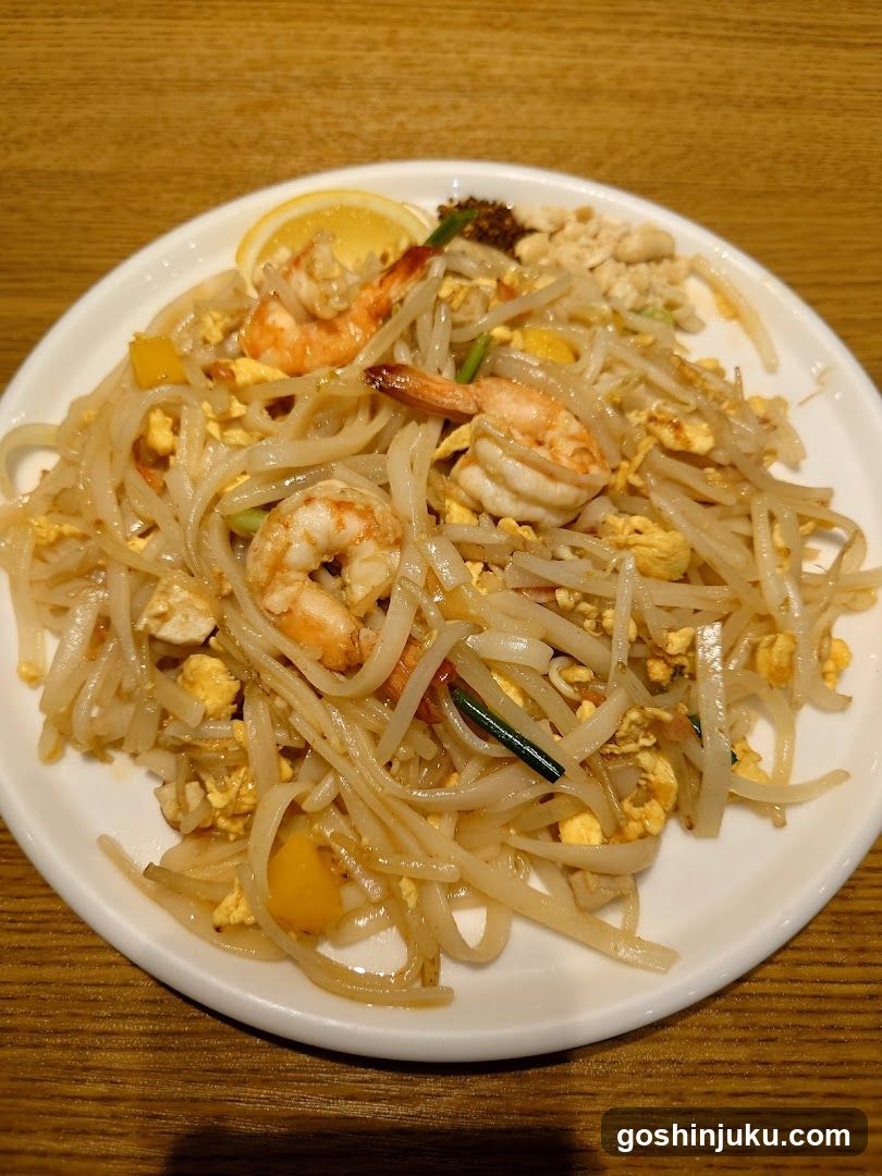 A plate of stir-fried rice noodles (pad thai) served on a white round dish, placed on a wooden table. The dish contains wide rice noodles, large shrimp, scrambled egg pieces, bean sprouts, green onions, and a lemon wedge garnish at the top edge.