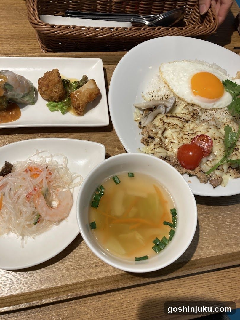 A table setting featuring a bowl of miso soup with green onions and carrots, a plate of glass noodles with shrimp, a rectangular dish containing spring rolls and fried items, and a rice bowl topped with grilled fish, mushrooms, cherry tomatoes, cilantro, and a sunny-side-up egg.