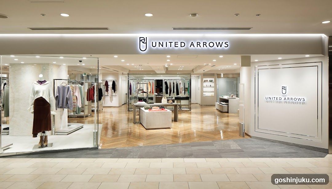 The image shows the exterior entrance of a United Arrows store, featuring illuminated signage with the brand logo and name above the glass facade. Inside, mannequins display clothing items on racks, with polished wooden flooring and bright lighting enhancing the retail environment.